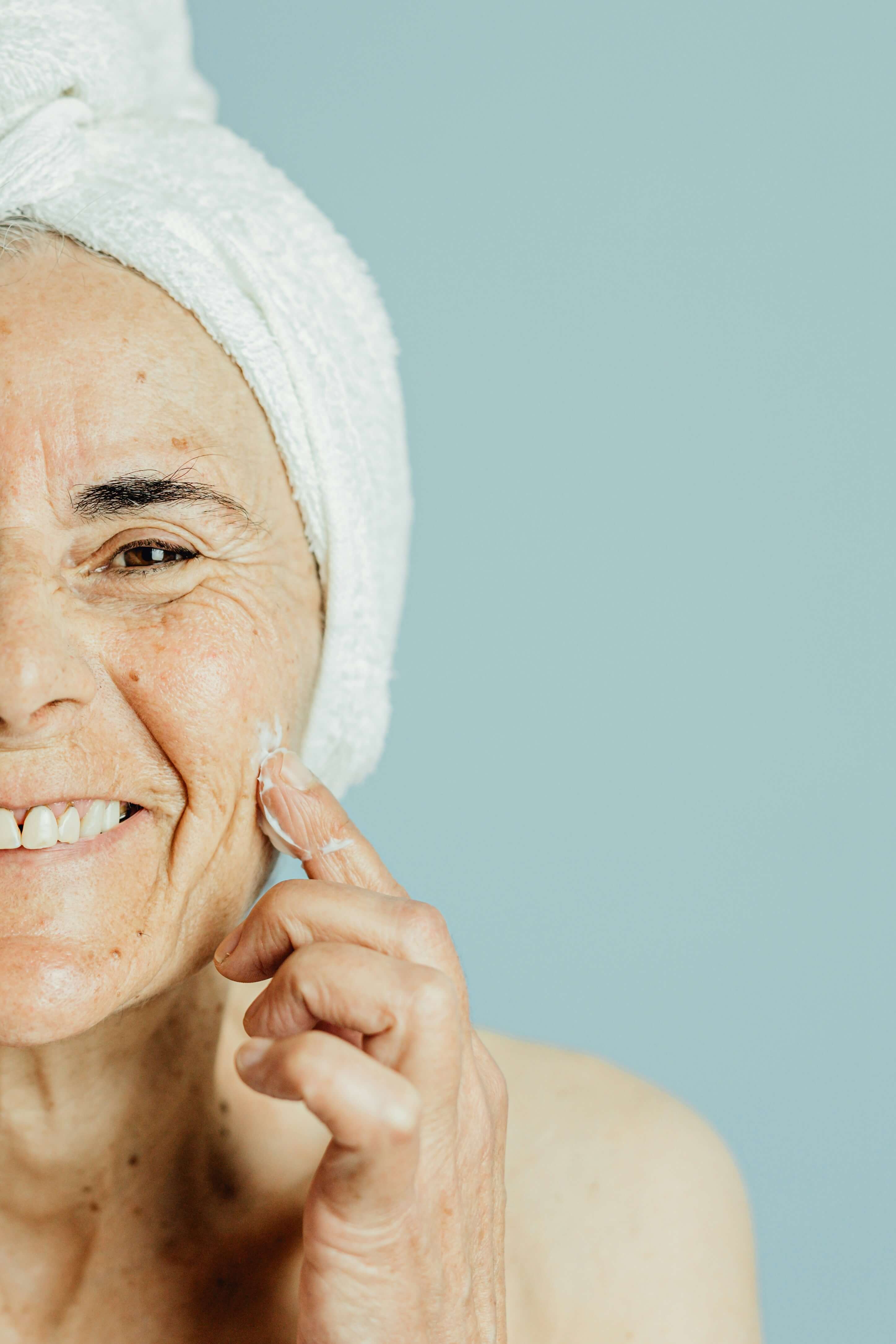 Older woman smiling at the camera with a towel round her head and putting on moisturiser