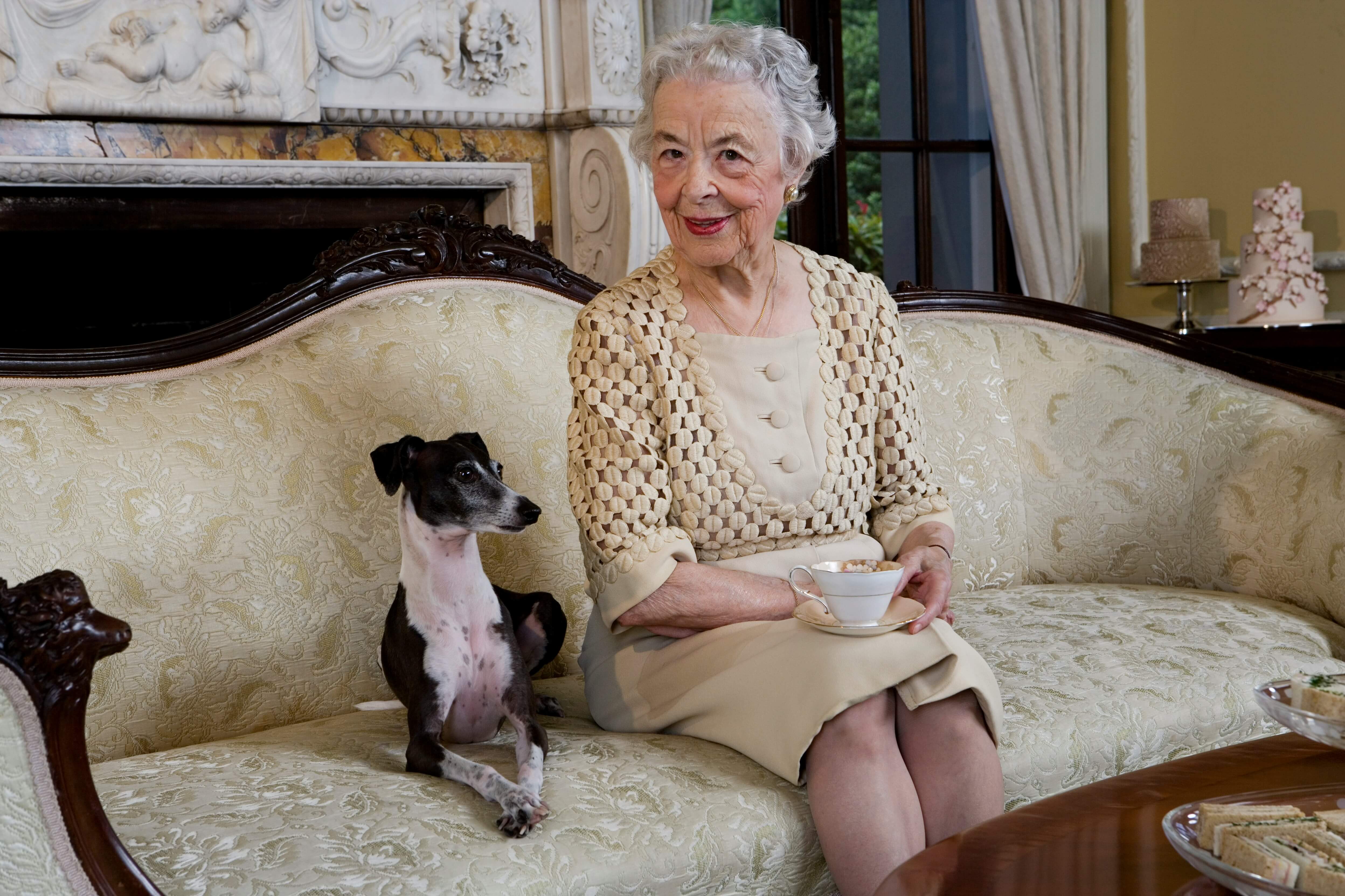 Older woman very prim drinking tea from a china cup and saucer wirth a small dog sitting next to her.